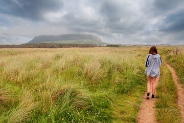 Naklejka na ściany i meble Young slim teenager girl walking on a small road towards Strandhill town, Knocknarea hill in cloudy sky in the background. County Sligo, Ireland. Irish landscape. Popular landmark.