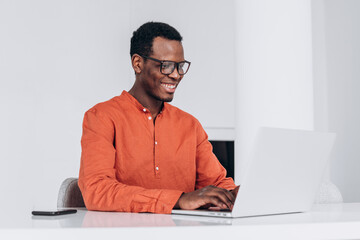 Concentrated young African-American guy in orange clothes and glasses works on modern laptop at...