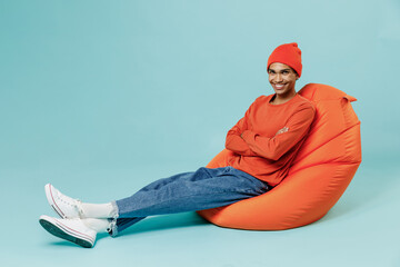 Full body young smiling happy african american man 20s wearing orange shirt hat sit in bag chair look camera isolated on plain pastel light blue background studio portrait. People lifestyle concept.