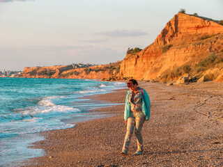 Brown-haired woman over 50 years old walks alone on beach. Woman with glasses and turquoise jacket....