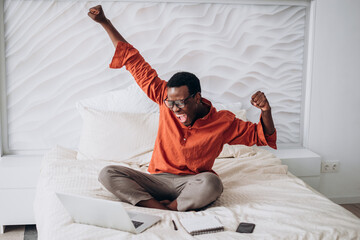 Joyful African-American guy in casual clothes and glasses puts hands up sitting near open laptop on comfortable bed at home