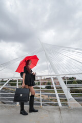White female business executive smiling with work briefcase, 40s long reddish brunette hair in black dress, wellies and open red umbrella on modern city bridge. Toledo, Spain