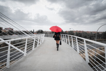 White female business executive smiling with business briefcase, 40s long reddish hair brunette with black dress, wellies and red umbrella walking with back to camera on modern city bridge. Toledo, Sp