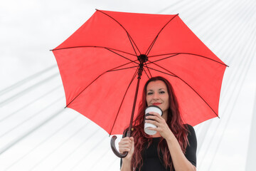 White female business executive smiling drinking coffee to go 40s long reddish brown hair brunette with black dress and red umbrella on modern city bridge. Toledo, Spain