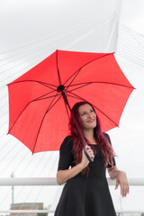 Smiling white female business executive in her 40s with long reddish brunette hair wearing black dress and red umbrella on modern city bridge. Toledo, Spain