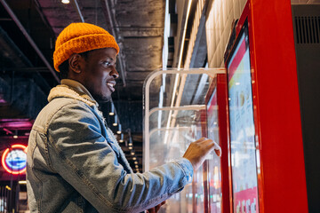 Positive African-American guy in orange knitted hat with wireless earphones orders food through...