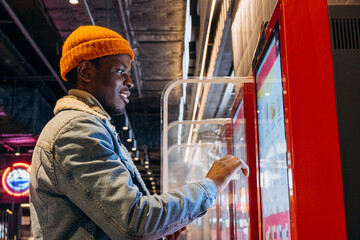 Positive African-American guy in orange knitted hat with wireless earphones orders food through self-service kiosk in cafe