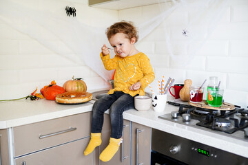Cute little girl sitting on the table near halloween decorations in the kitchen and eating pie with pumpkin
