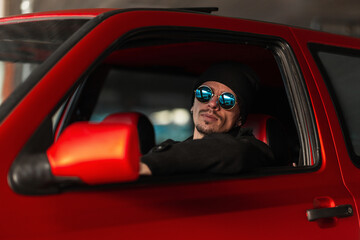 Fashionable cool young hipster man in blue sunglasses and black hat is driving a red car