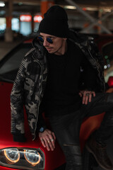 Fototapeta na wymiar Stylish cool handsome model man with vintage sunglasses in a fashionable military jacket with a black hat and pullover sits near a red car on the street