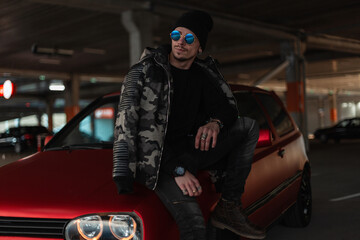 Fototapeta na wymiar Fashion urban man model with sunglasses and hat in stylish winter military jacket and pullover stands near a red car at parking lot