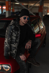 Fototapeta na wymiar Stylish hipster man with fashion sunglasses in urban military jacket, pullover, jeans and hat stands near red car in the city