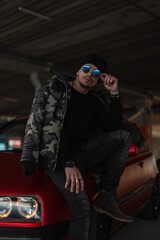 Fototapeta na wymiar Fashion man with stylish sunglasses in winter military jacket, black pullover and hat poses near red car on the street