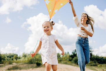 A laughing little girl and her mother run along the road to the field and fly a kite. Mom and daughter have fun together