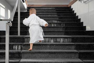 Back view of cute funny caucasian blond kid boy wearing white pool spa bathrobe walking upstairs on...
