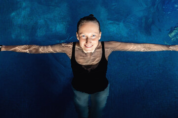 Above top view portrait of young adult caucasian female model in sport black swimsuit stand in clear blue water of indoor swimming pool at dark evening time. Relax harmony and wellness spa therapy