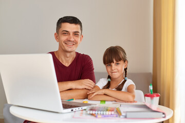 Indoor shot of smiling handsome man sitting at table with his little daughter in front of laptop, looking at camera with happy expression, distant education.