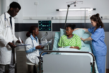 Multi ethnic medical team consulting young person in hospital ward bed at clinic. Caucasian nurse and african american doctors talking to patient of african ethnicity about illness, sickness