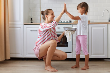 Side view photo of cheerful charming mommy and small kid giving high-five in light kitchen, baking...