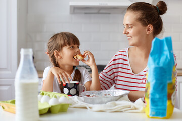 Portrait of young woman and her little cute daughter sitting at table and eating cakes and cookies on kitchen and drinking beverage, having fun together while enjoying freshly baked pastries.