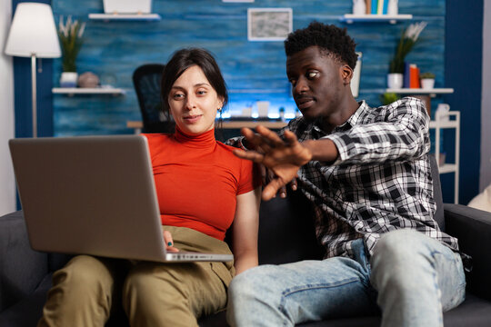 Married Interracial Couple Looking At Modern Laptop On Couch. Multi Ethnic Partners Using Digital Device With Technology, Watching Display Of Gadget. Mixed Race People With Computer