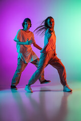 Fototapeta na wymiar Young hip-hop dancers, stylish emotive girl and boy in action and motion in casual sports youth clothes on gradient multi colored background at dance hall in neon light.