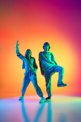 Fototapeta na wymiar Young dancers, stylish emotive girl and boy dancing hip-hop in casual sports youth clothes on gradient multi colored background at dance hall in neon light.