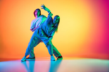 Fototapeta na wymiar Young people, hip-hop dancers, stylish emotive girl and boy in action and motion in casual sports youth clothes on gradient multi colored background at dance hall in neon light.