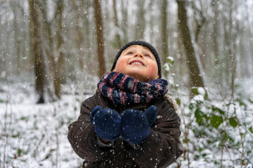 Close up portrait of a little boy playing with snowflakes in a park in winter. Happy child enjoys the first snow in a forest.