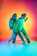 Rolgordijnen Young dancers, stylish emotive girl and boy dancing hip-hop in casual sports youth clothes on gradient multi colored background at dance hall in neon light. © master1305