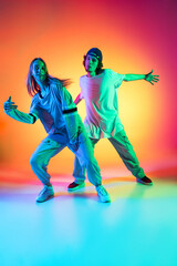 Fototapeta na wymiar Young hip-hop dancers, stylish emotive girl and boy in action and motion in casual sports youth clothes on gradient multi colored background at dance hall in neon light.