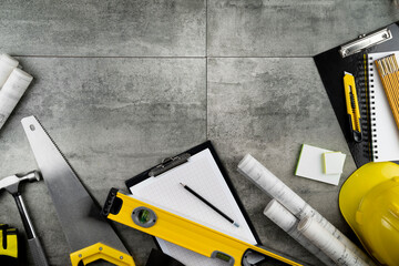 Contractor concept. Tool kit of the contractor: yellow hardhat, libella, hand saw. Plans and notebook on the gray tiles background.