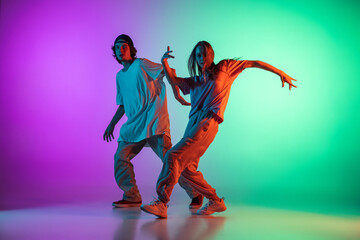 Young hip-hop dancers, stylish emotive girl and boy in action and motion in casual sports youth clothes on gradient multi colored background at dance hall in neon light.