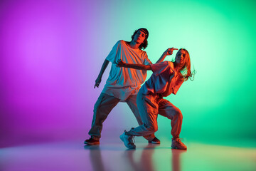 Fototapeta na wymiar Stylish young hip-hop dancers, emotive girl and boy in action and motion in casual sports youth clothes on gradient multi colored background at dance hall in neon light.