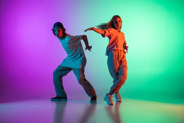 Fotobehang Stylish young hip-hop dancers, emotive girl and boy in action and motion in casual sports youth clothes on gradient multi colored background at dance hall in neon light. © master1305