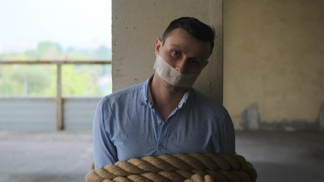 businessman tied with rope with his mouth sealed with tape sits tied to post, looks up at camera. Seen man in blue shirt, victim of maniac with gag in his mouth, cannot speak, dumb.