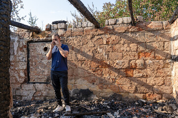 Young man calls by mobile phone, standing in house destroyed with fire, tragic face, crying. Damaged window, walls and floor, burnt wooden frame, all ruins. Sun shines from holes in roof. Insurance.