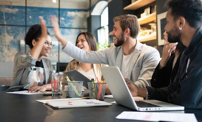 Two cheerful young business people giving high-five while their colleagues looking at them and...