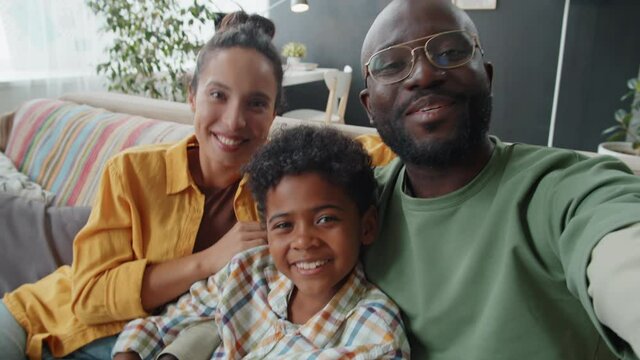 Happy Afro-American parents and little son sitting on sofa at home, waving, smiling and speaking on camera via online video call