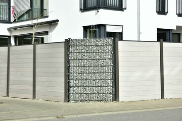 Moderner Sichtschutzzaun abwechselnd aus mit Naturstein gefüllten Gabionen aus verzinktem Stahlgitter und Kunststoffverschalung um ein bebautes Grundstück mit Wohnkomplex © Hermann