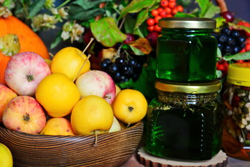 Autumn still life. Wooden bowl of colorful apples and jars of homemade herbal and nuts honey close up on background of wicker basket with bunch of red and black ash berries, pumpkin and branch of hops