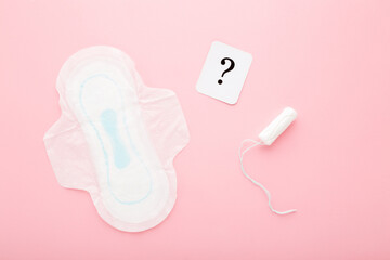 Card of question mark, new white sanitary towel and tampon on light pink table background. Pastel color. Closeup. Choice of female hygiene products. Top down view.