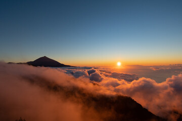 Views of a sunset with the Teide volcano above the clouds and the sun hiding between them.