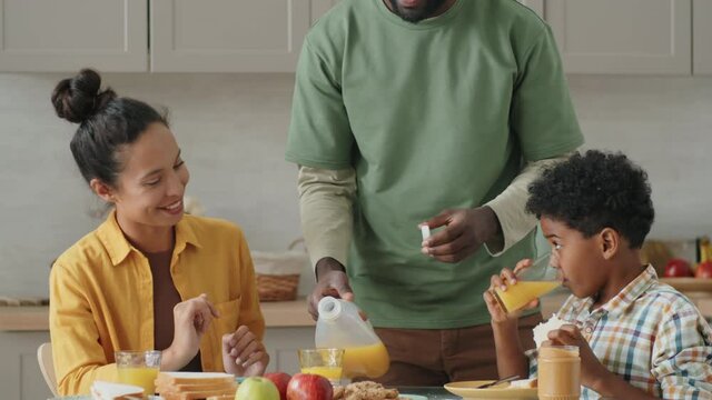 Afro-American man pouring orange juice into glass for wife, then sitting at kitchen table and talking with family during breakfast at home