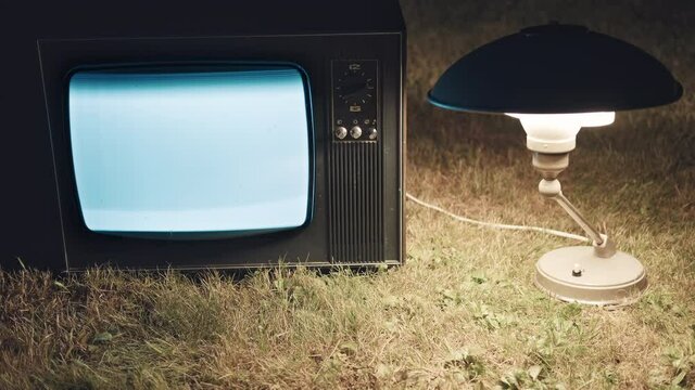 Old retro TV is standing on the grass. There is a vintage lamp nearby. Concept of surrealism, horror and old school vintage technique of the last century