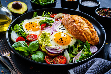 Sunny side up eggs on bun with  fresh green vegetables, mini tomatoes and guacamole on wooden table
