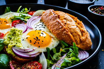 Sunny side up eggs on bun with  fresh green vegetables, mini tomatoes and guacamole on wooden table
