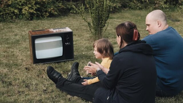Family of a man, a woman and little baby are sitting on the grass and watching an old retro TV. They clap their hands. TV is strobing. The concept of post apocalypse, horror. Dependence on television
