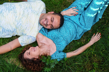 Loving couple lie on the grass look at each other and laugh. Man in a T-shirt and a curly woman in a blue dress on a romantic date. A Middle Eastern man with his caucasian wife.