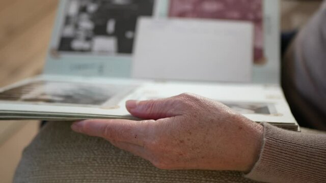 Adult woman looks old photo album at home. Close up view of aged woman's hand with old photo album. Elderly person is leafing through photo album.